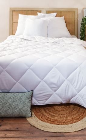 Chambre moderne avec lit à draps blancs, tête de lit tressée, table de nuit en bois décorée, tapis géométriques sur parquet, et coussin vert – décoration intérieure naturelle et cosy