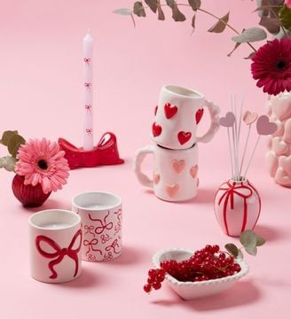 Tasses blanches d&eacute;cor&eacute;es de c&oelig;urs rouges et d&rsquo;un n&oelig;ud, pos&eacute;es sur un fond rose avec fleurs et petits objets d&eacute;coratifs.